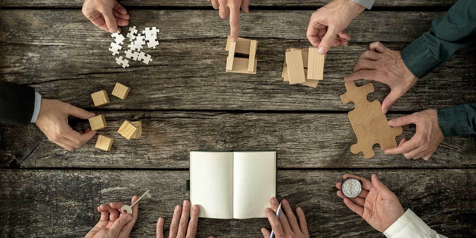 Photograph of eight people's hands in a circle working with various objects such as puzzle pieces, stacks of cubes, and a notebook and pencil