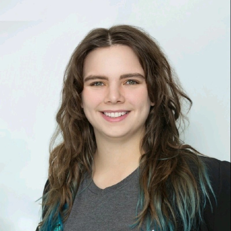 Professional headshot of a smiling woman with long brown hair, wearing a gray shirt and dark blazer against a light background.