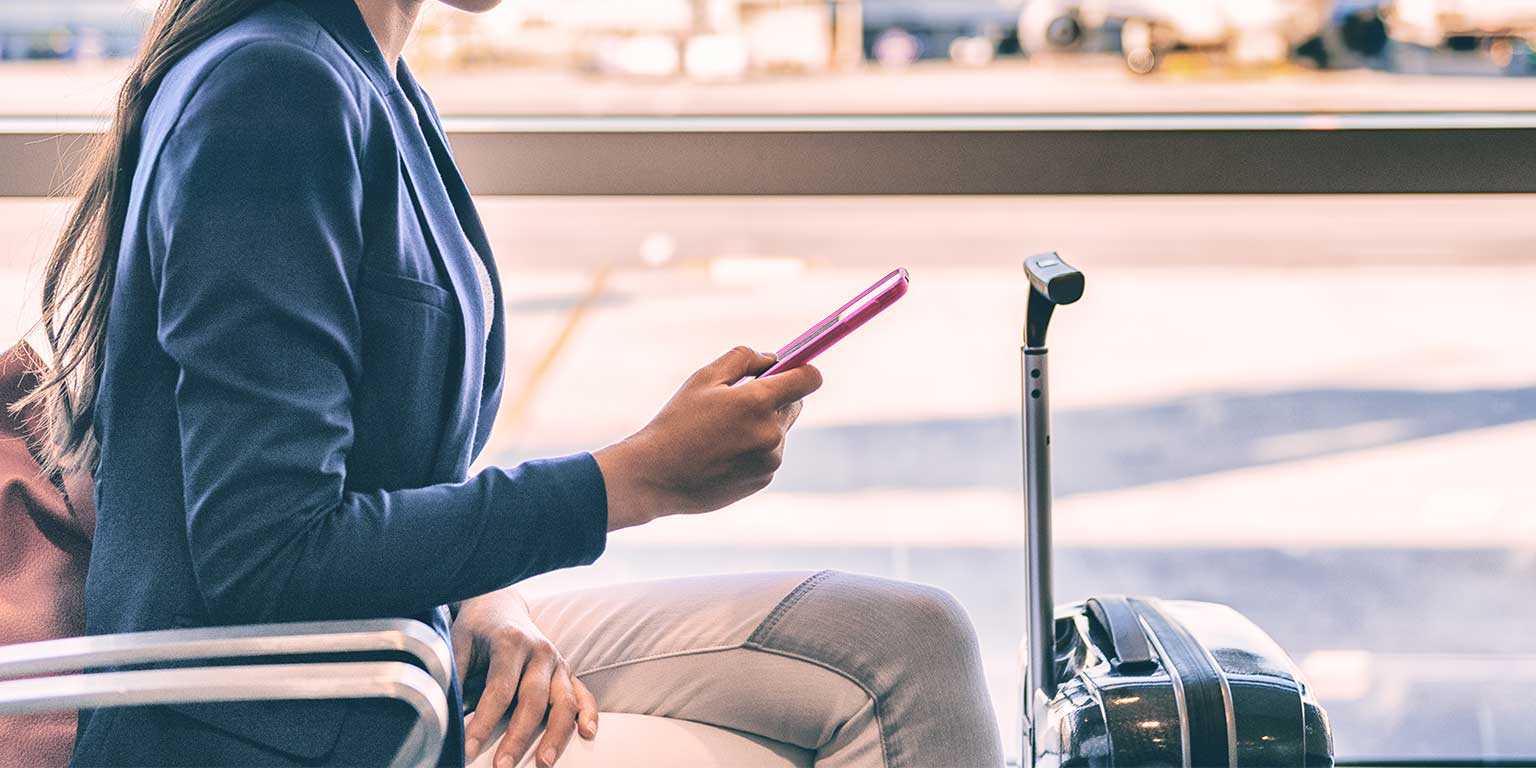 Photograph of a sales rep looking at their phone and sitting at the airport with a suitcase